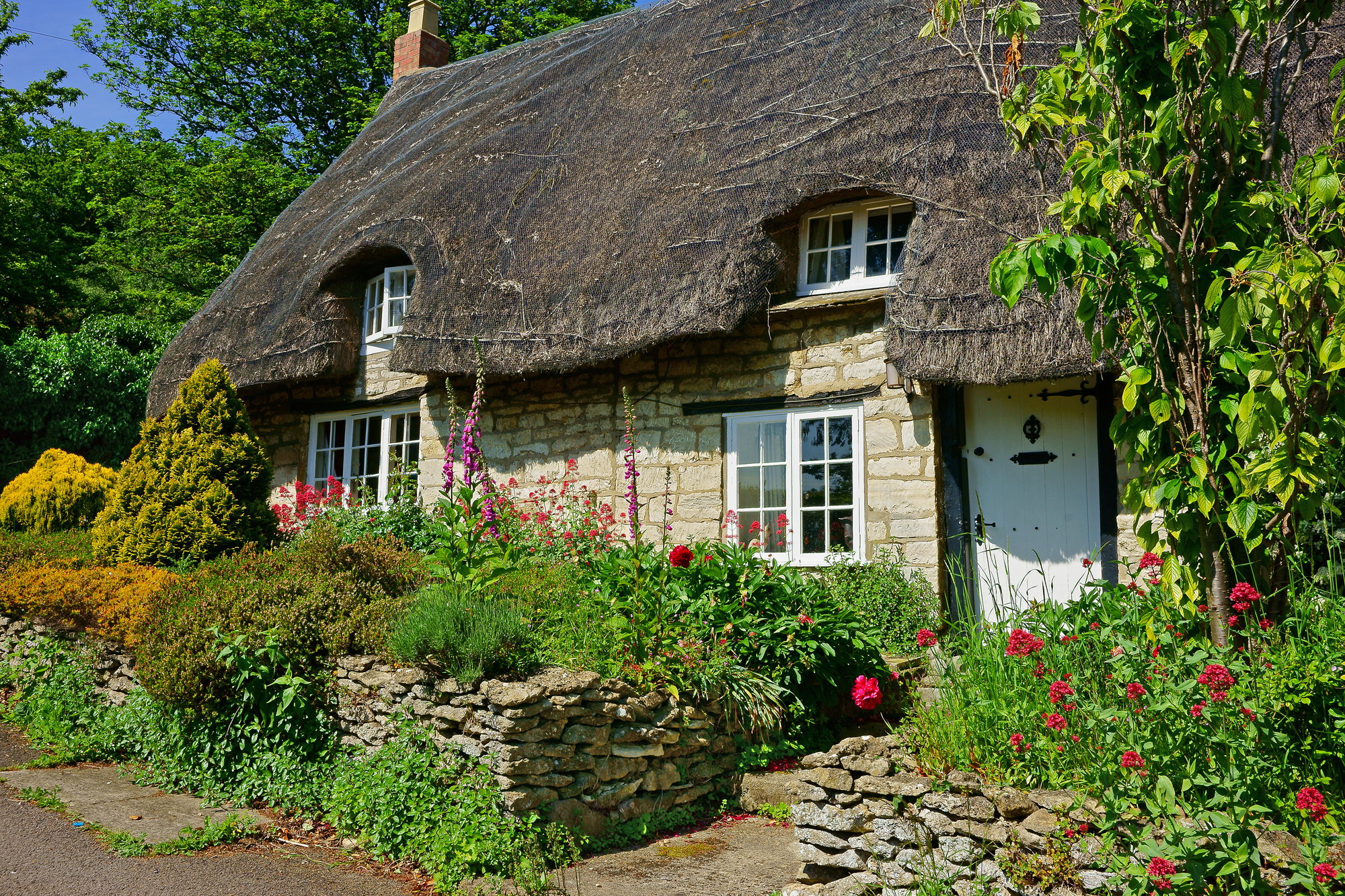 An image of a quaint and charming Cotswold cottage with a thatched roof and flowering garden during the summer