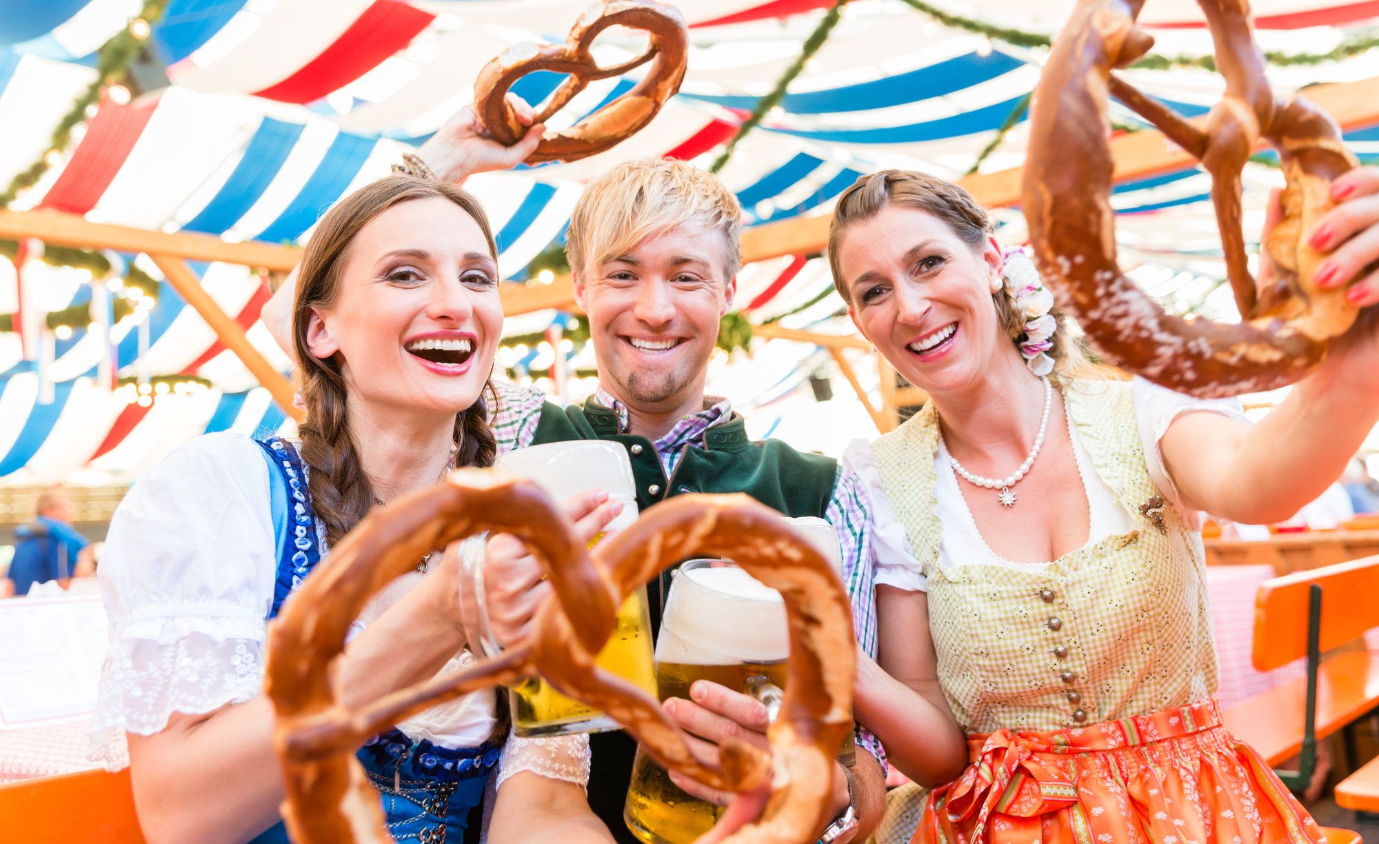 Three friends dressed in traditional Bavarian clothing hold up steins of beer and giant pretzels in a tent during Oktoberfest in Munich, Germany